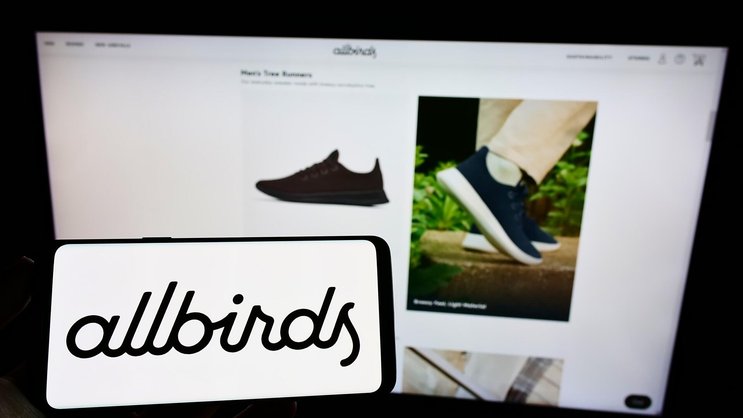 Person, die ein Mobiltelefon mit dem Logo des Schuhherstellers Allbirds auf dem Bildschirm vor einer Unternehmenswebseite hält.