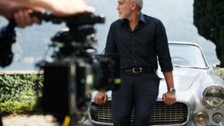 George Clooney ist einen silberfarbenen Maserati 3500 GT Spyder Vignale angelehnt und schaut auf die Seite.