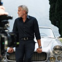 George Clooney ist einen silberfarbenen Maserati 3500 GT Spyder Vignale angelehnt und schaut auf die Seite.
