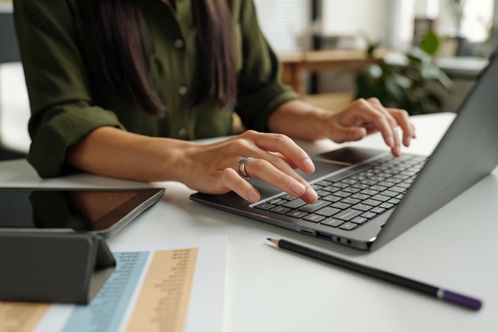 Hand of young female teacher or student typing on laptop keyboard while sitting by desk in front of camera and answering task questions